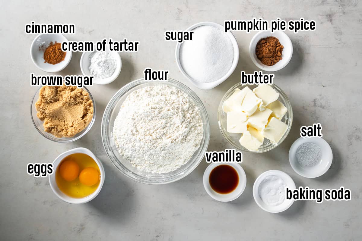 Ingredients for Pumpkin Spice Snickerdoodles arranged in labeled bowls, including flour, butter, sugars, eggs, vanilla, cream of tartar, baking soda, salt, and spices.