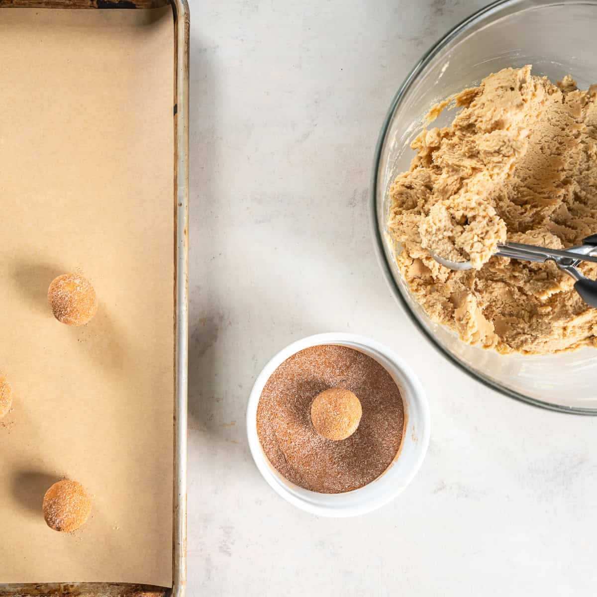 A ball of snickerdoodle dough in spiced sugar alongside a bowl of dough and a parchment-lined baking sheet.