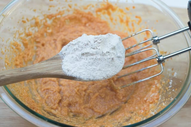 A spoon adds dry mixture to a wet mixture in a glass bowl.