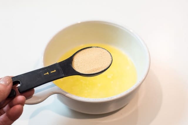 A hand holding a measuring spoon filled with garlic powder over a bowl of melted butter and oil.