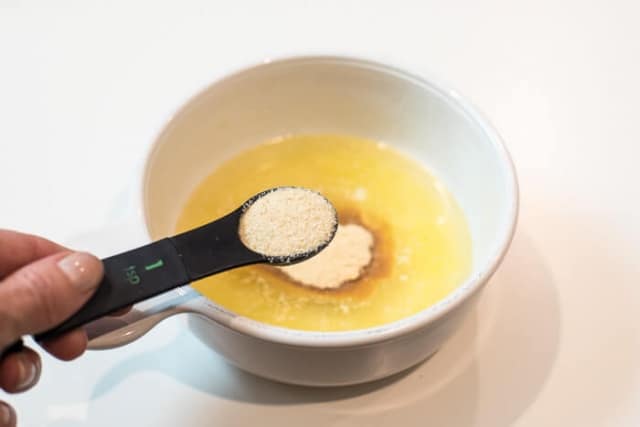 A hand holding a measuring spoon filled with onion powder over a bowl of melted butter.