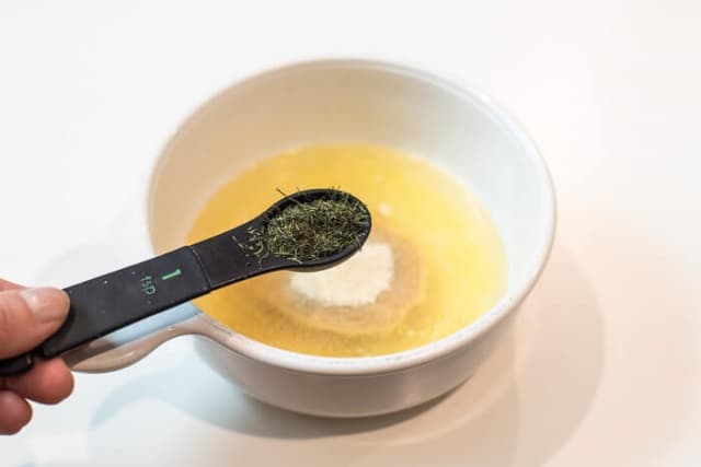 A hand holding a measuring spoon filled with dried dill weed over a mixing bowl filled with melted butter and seasoning.
