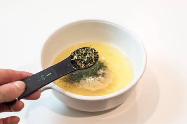 A hand holding a measuring spoon filled with dried parsley over a bowl with melted butter and seasoning in it.