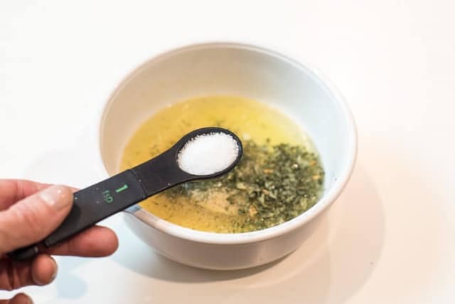 A hand holding a measuring spoon filled with salt over a bowl with melted butter and seasoning in it.