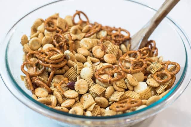 A wooden spoon stirring Garlic Dill Snack Mix in a glass mixing bowl.