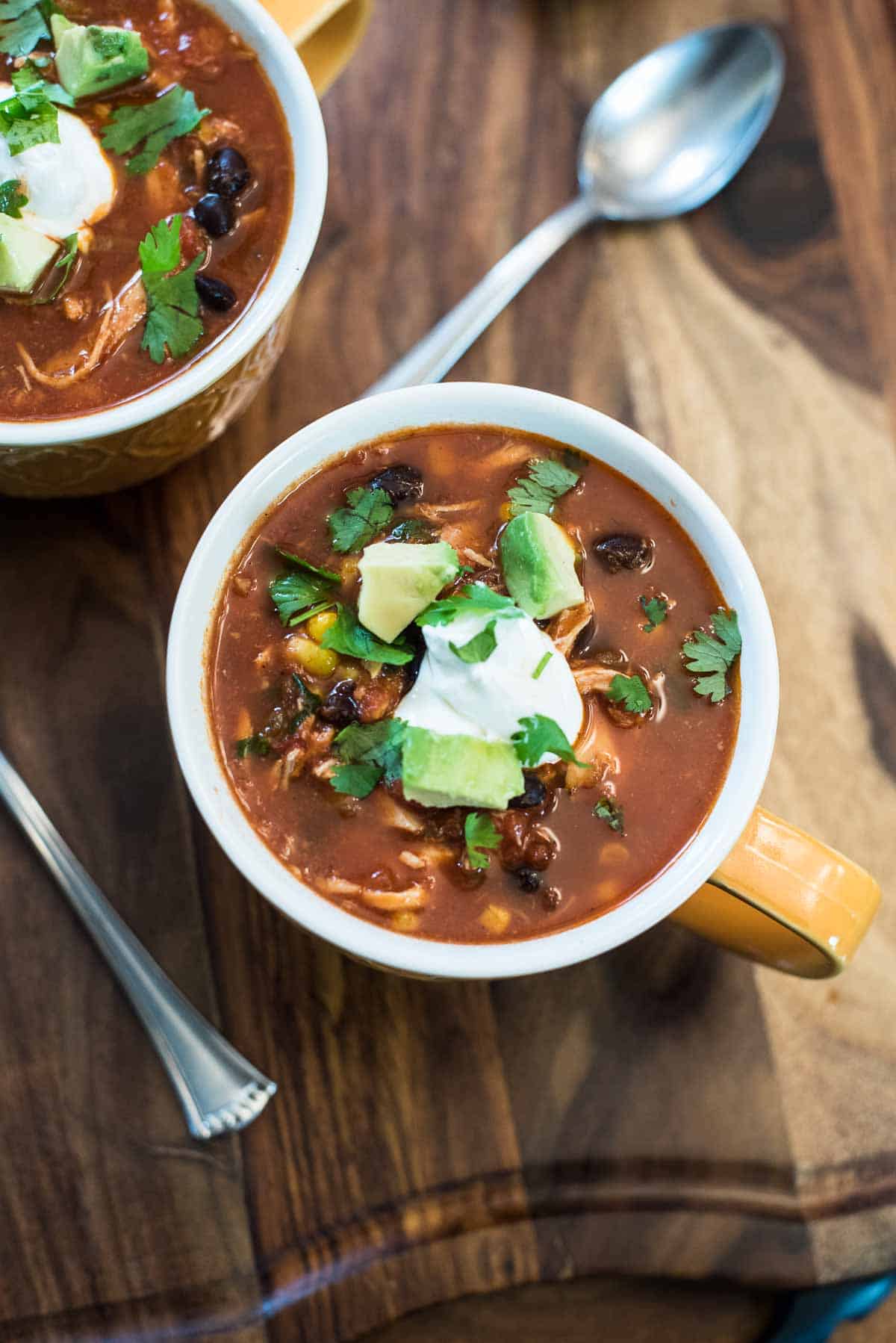 Two mugs of chicken tortilla soup topped with sour cream and cilantro on a wood board.