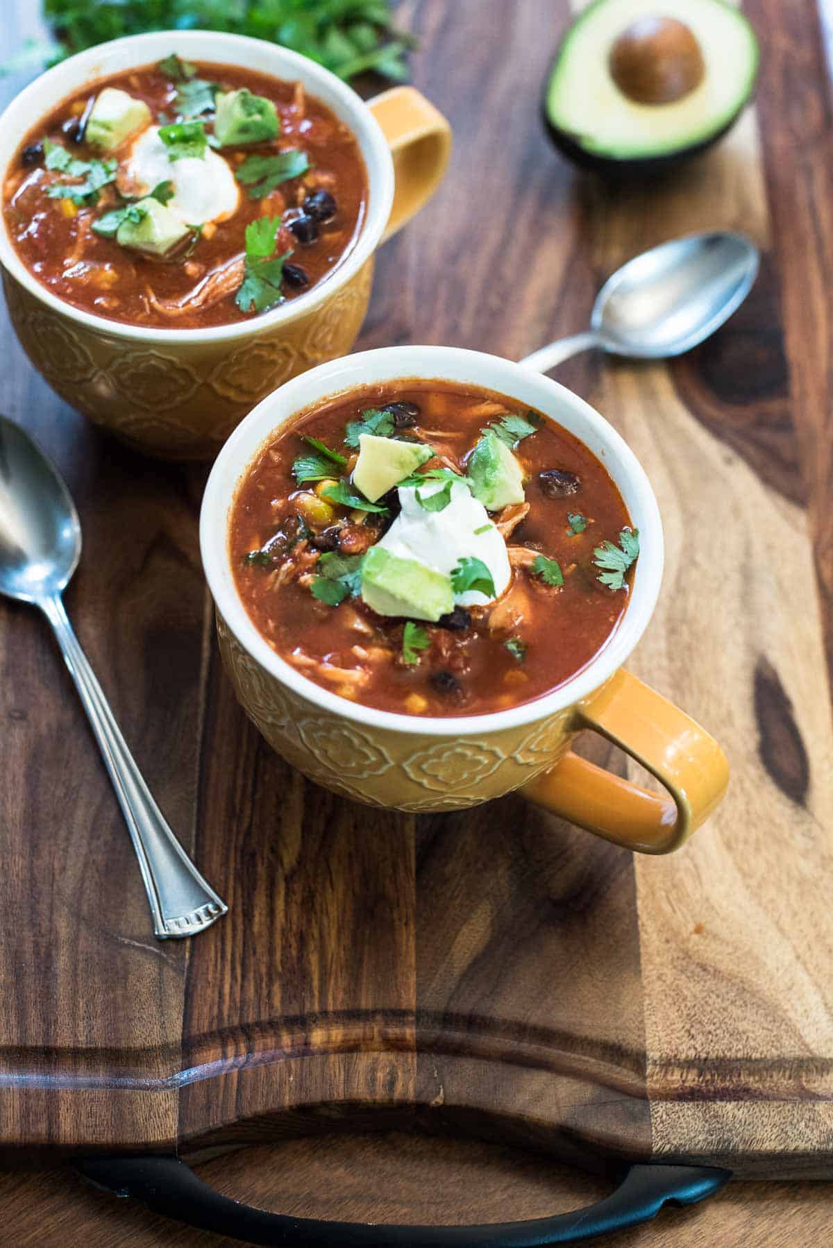 Two mugs of chicken tortilla soup and spoons resting on a wood board.