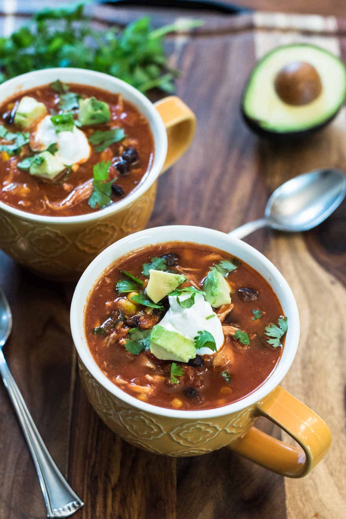 Two mugs of chicken tortilla soup topped with sour cream on a wood board.