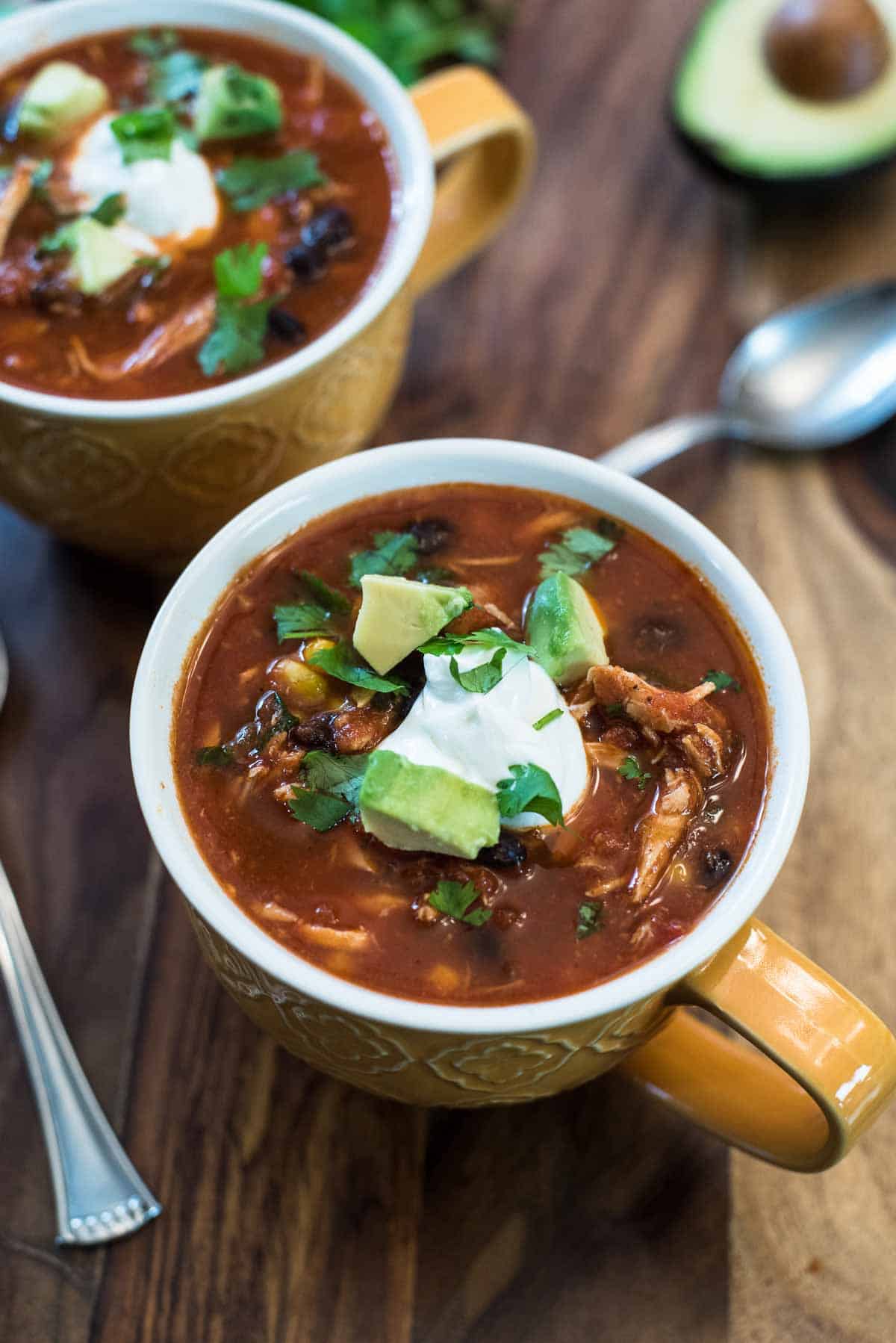 Two mugs of chicken tortilla soup topped with sour cream on a wood board.