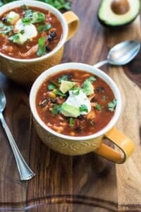 Two mugs of chicken tortilla soup topped with sour cream on a wood board.