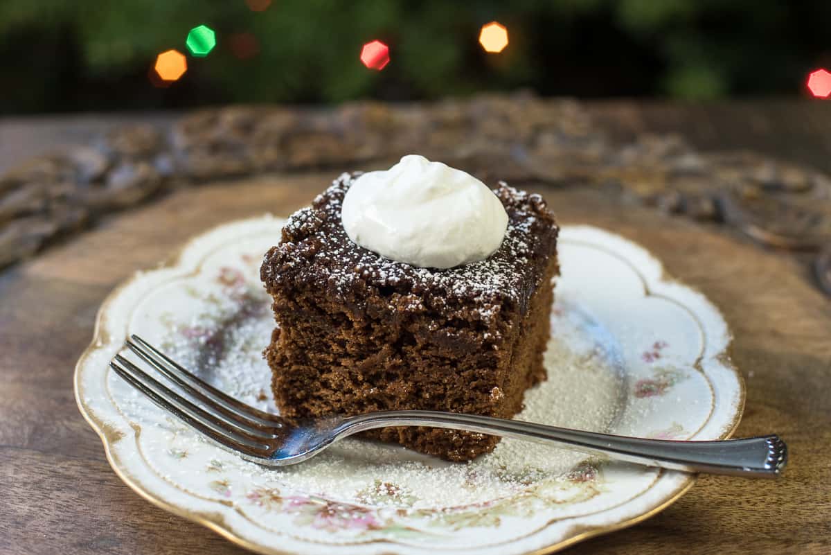 A slice of gingerbread topped with powdered sugar and whipped cream on a pretty china plate with a fork.