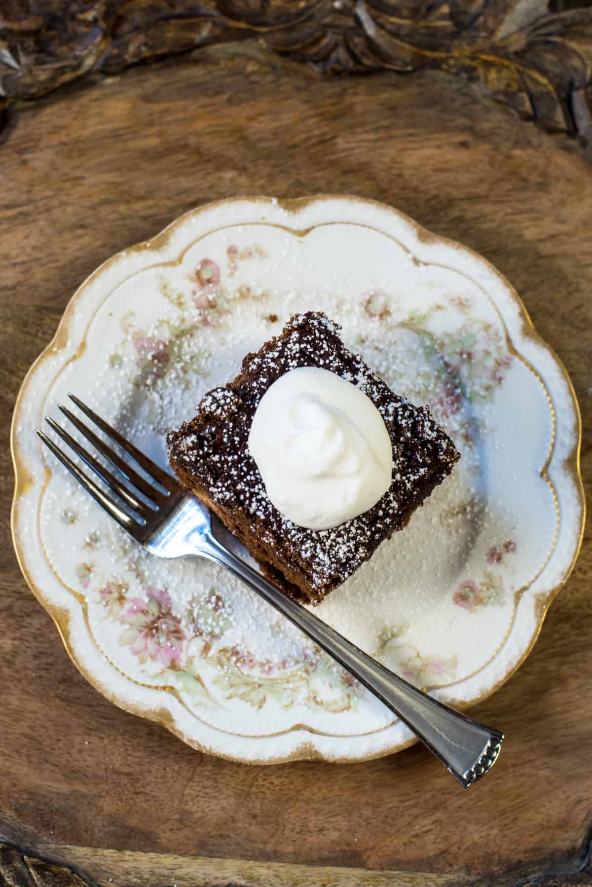A top down shot of a slice of gingerbread topped with whipped cream on a pretty china plate with a fork.