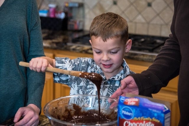 Brownie batter pours off a spoon held by a smiling young boy into a mixing bowl.