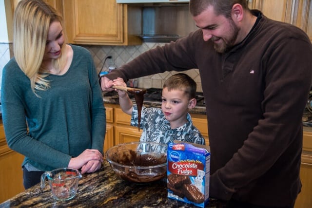 A young man helps a boy lift a wooden spoon up from a mixing bowl as a young woman watches next to them.