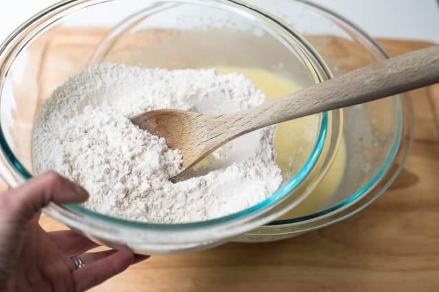 A hand holds a bowl of dry ingredients and a wooden spoon.