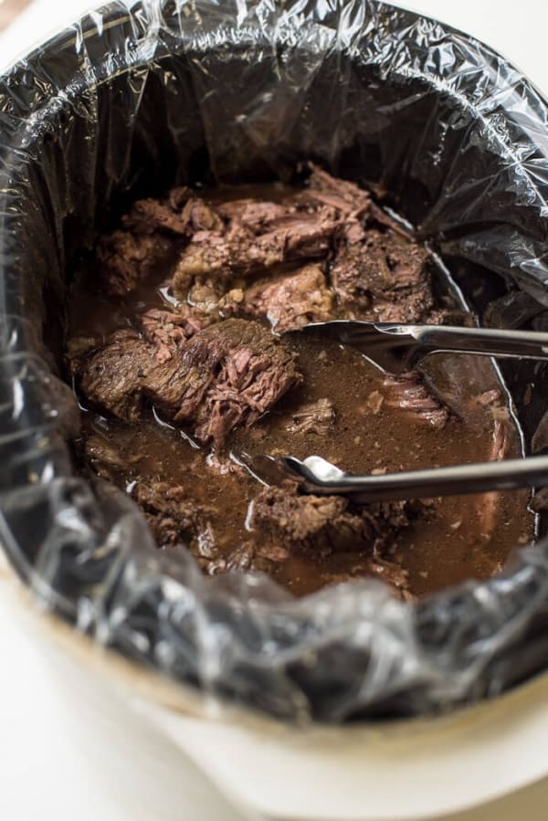 Metal tongs in a crock pot with cooked beef.