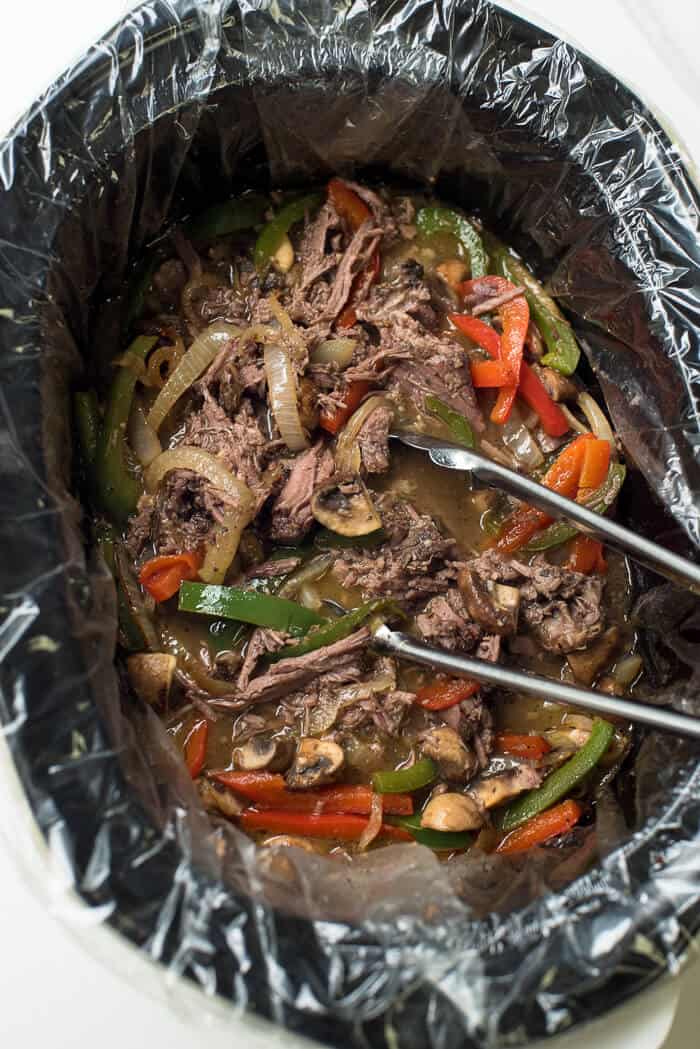 Cooked beef and vegetables in a crock pot with metal tongs.