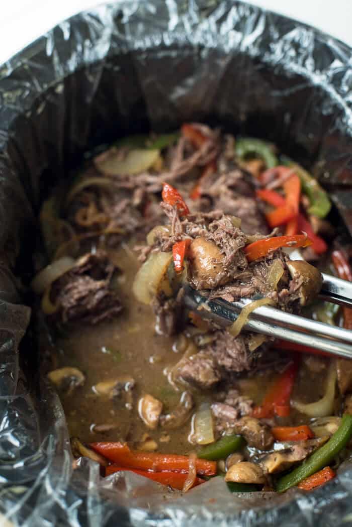 Tongs lift cooked beef and vegetables from a slow cooker.