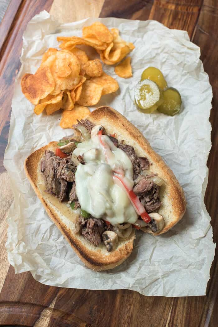 A sandwich with melted cheese on parchment paper with chips and pickles.