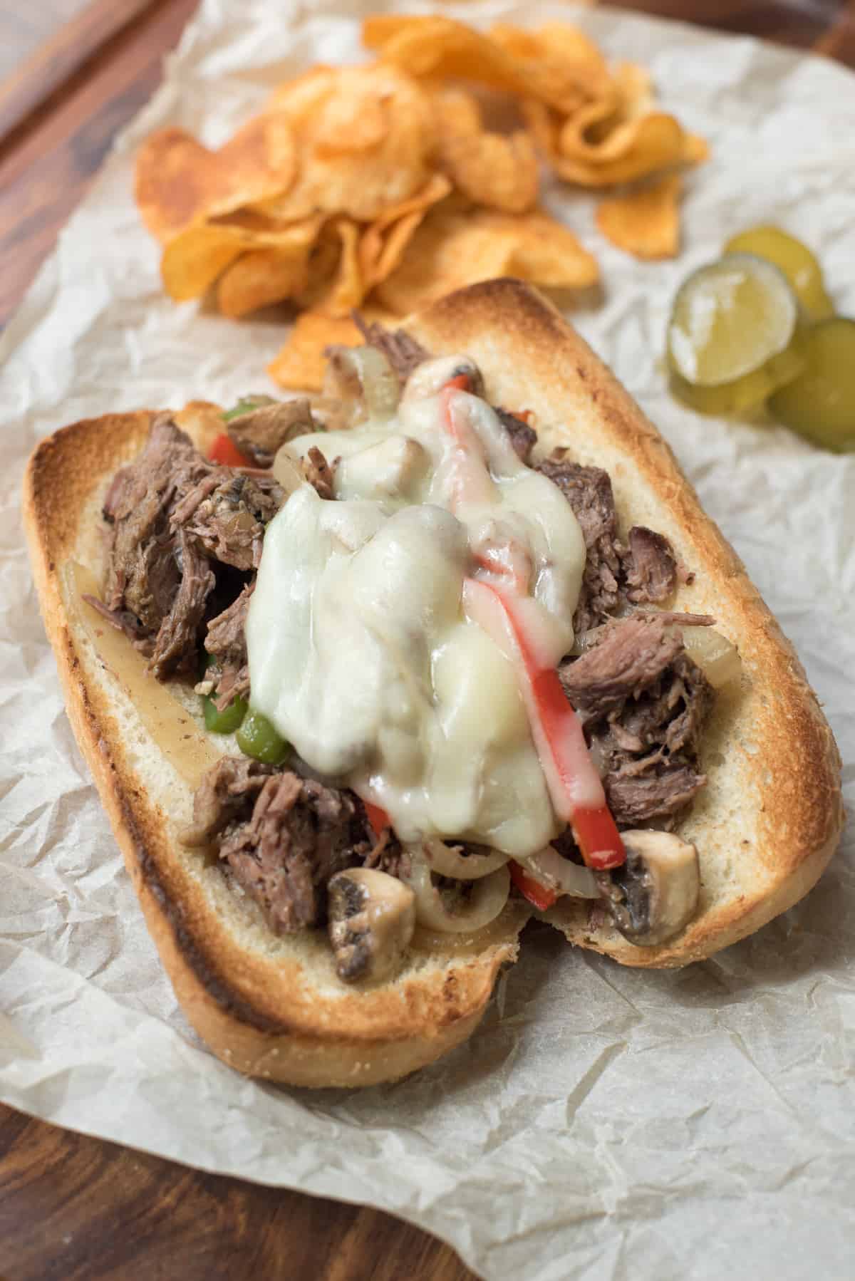 A drip beef sandwich on parchment paper next to potato chips and pickles.