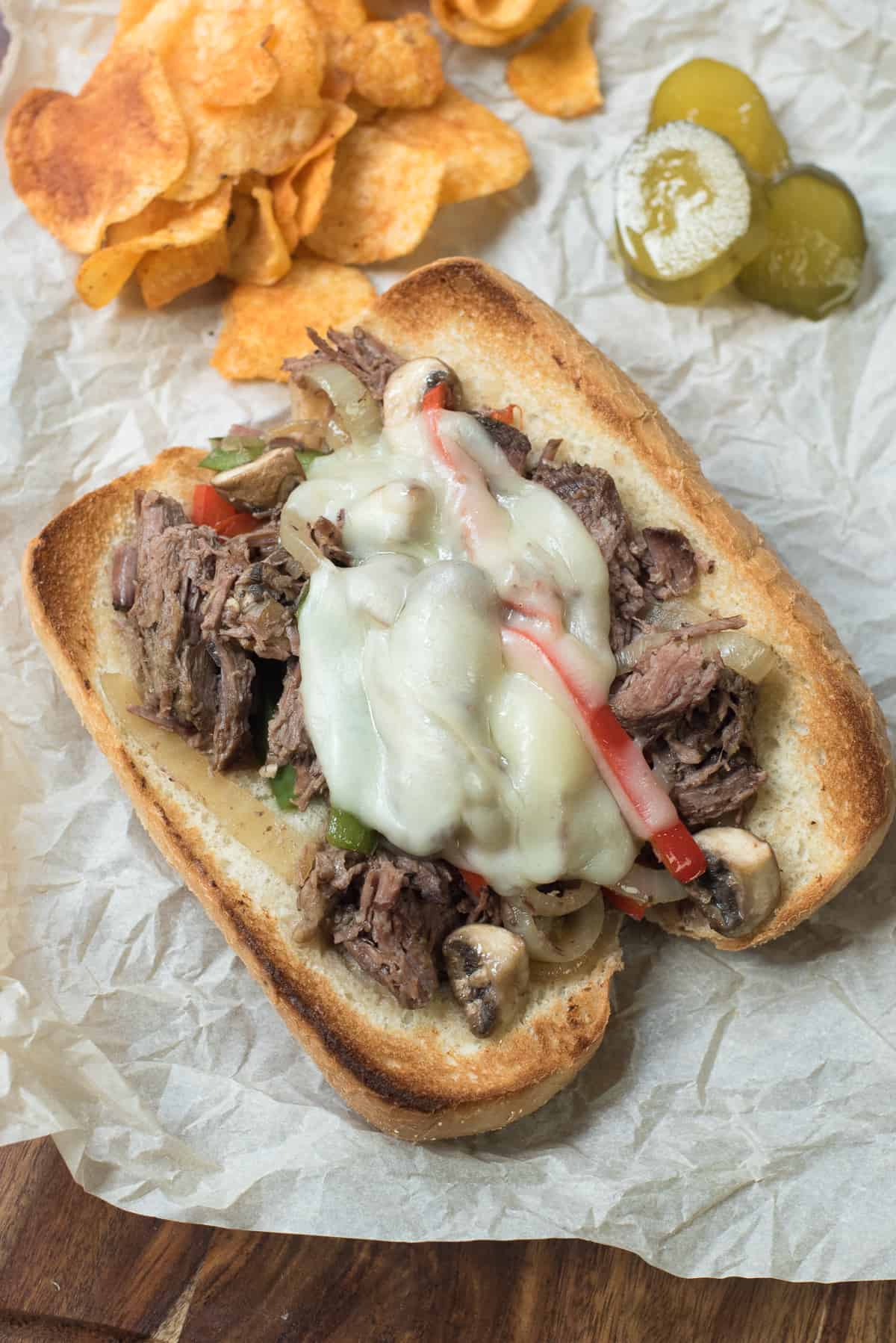 A drip beef sandwich on parchment paper next to potato chips and pickles.