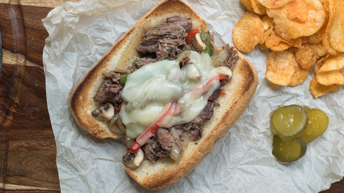 A drip beef sandwich on parchment paper next to potato chips and pickles.