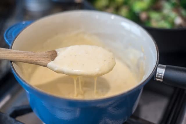 A spoon scoops up a cheesy white sauce from a saucepan.