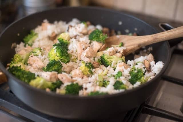 A spoon stirs, rice, broccoli, and mushrooms in a skillet on the stove.