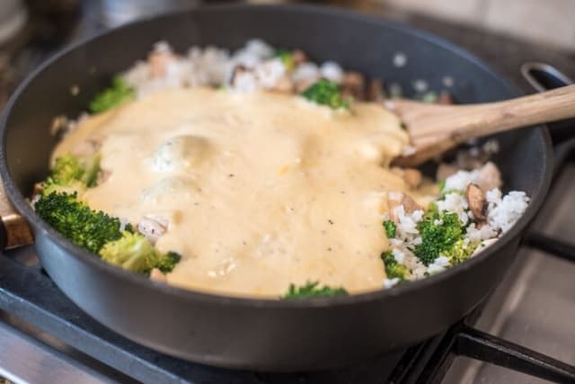 A cheesy white sauce on top of broccoli, rice, and mushrooms in a skillet on the stove.