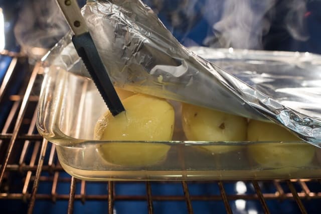 A knife piercing a peeled baked potato in a baking dish in the oven.