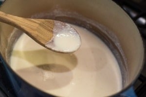 A thickened white sauce dripping from a wooden spoon into a Dutch oven.