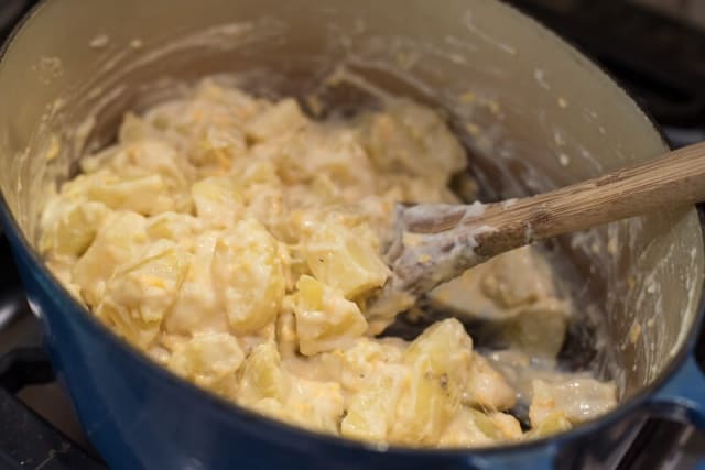 A wooden spoon stirs cooked potatoes into a cheese sauce in a Dutch oven.