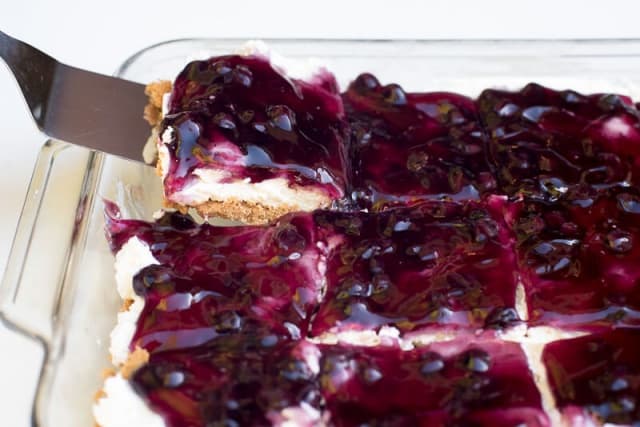 A spatula lifts a piece of blueberry cheesecake dessert from a baking dish.
