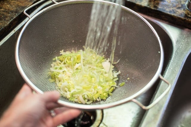 Sliced leeks in a colander being rinsed with water from a faucet.