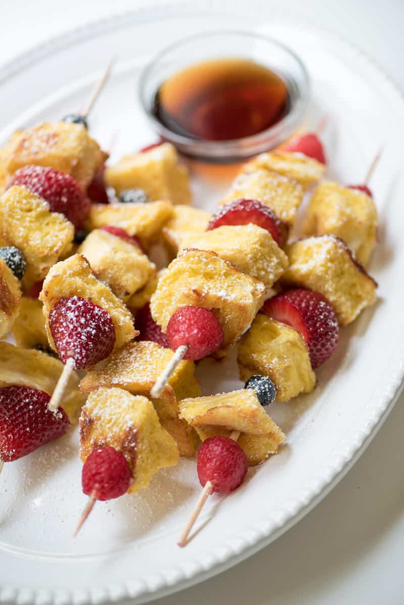 Berry French Toast Kabobs stacked on a white serving plate with a small bowl of maple syrup.