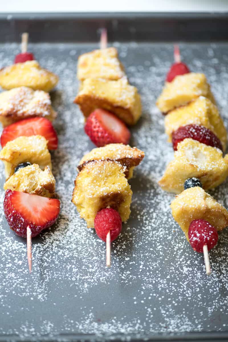 Cubes of French toast and berries on wooden skewers dusted with powdered sugar on a baking sheet.