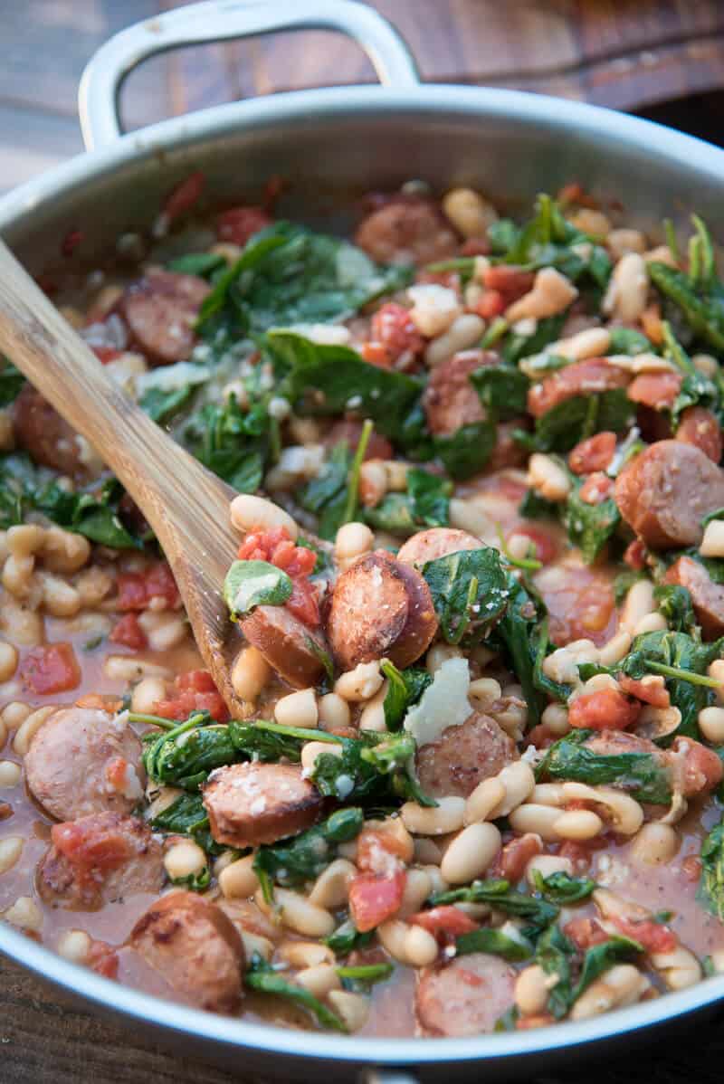 Skillet Sausage and White Beans with Spinach is stirred in a skillet with a wooden spoon.