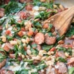 A close up of a wooden spoon in a skillet with sausage, white beans, and spinach.