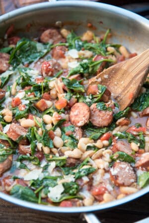 A close up of a wooden spoon in a skillet with sausage, white beans, and spinach.