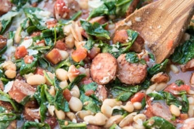 A close up of a wooden spoon in a skillet with sausage, white beans, and spinach.