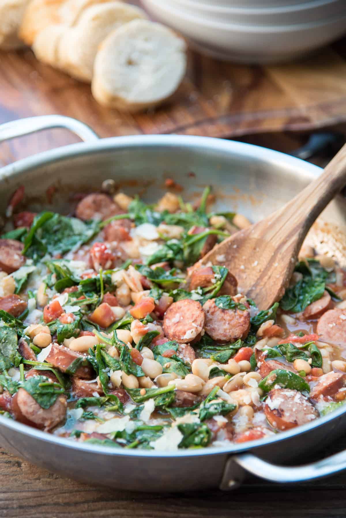 A wooden spoon in a skillet with sausage, white beans, and spinach.