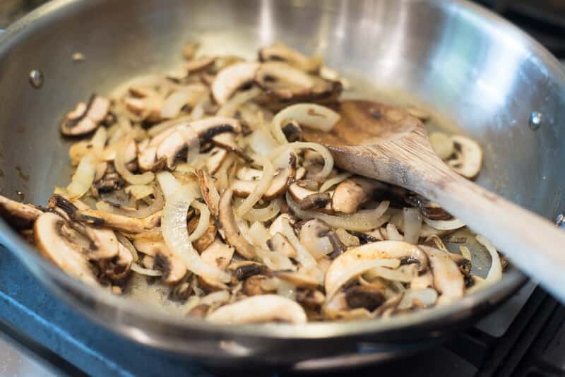 Mushrooms and onions in a skillet with a wooden spoon.