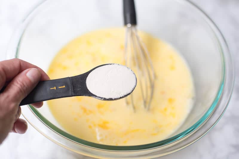 A measuring spoon full of flour held over a bowl of whisked eggs and milk.