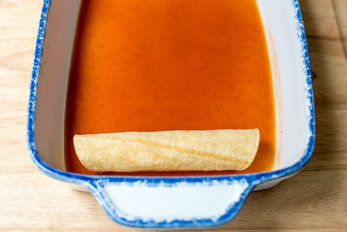 A white casserole dish coated with enchilada sauce with one filled and rolled tortilla.