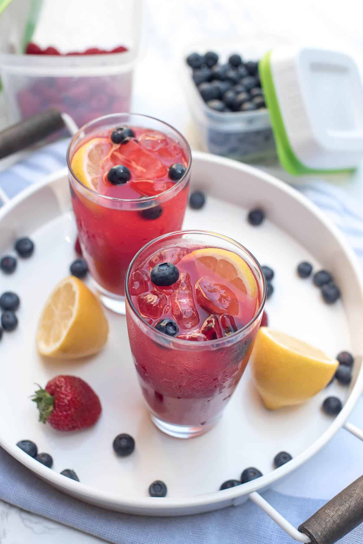 An overhead shot of two glasses of Berry Lemonade on a white tray with lemons, blueberries, and strawberries.