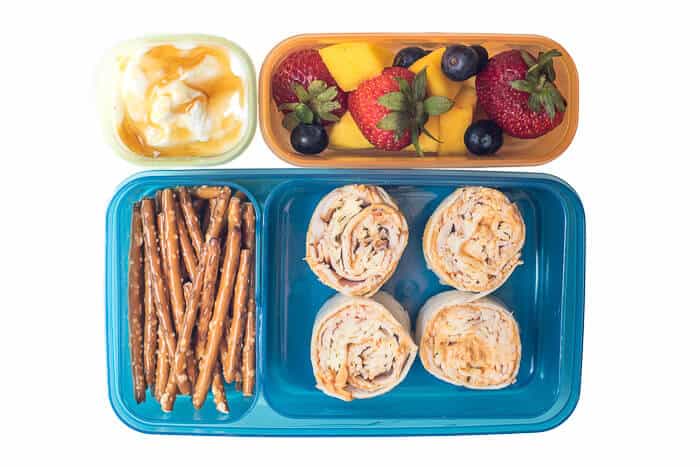 Tortilla pinwheels, pretzels, fruit, yogurt and honey in a plastic lunch container.