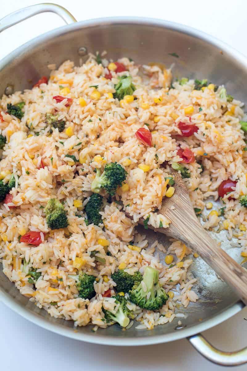 A wooden spoon scooping. Cheesy Vegetable Rice from a skillet.