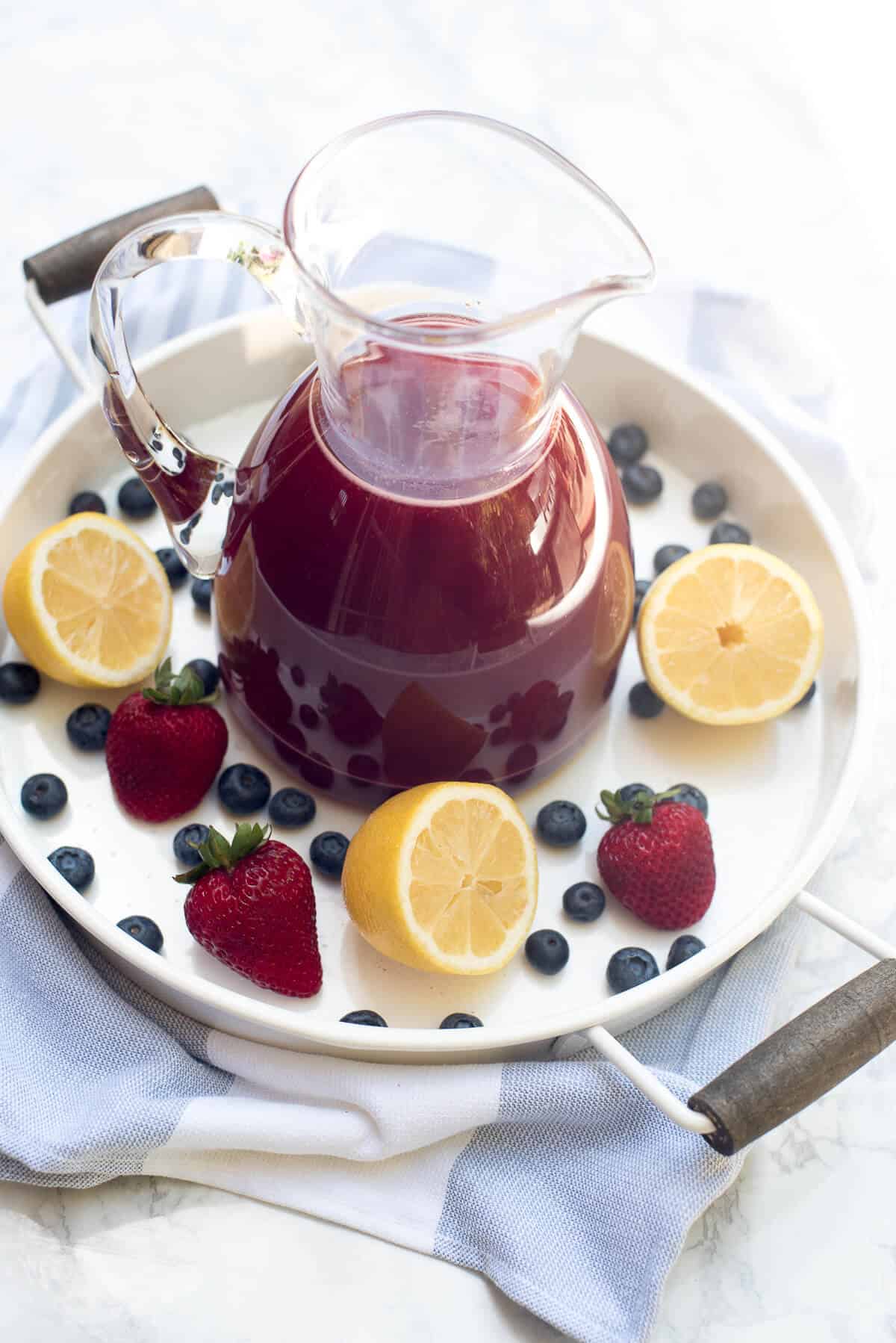 A pitcher of Berry Lemonade on a white tray with lemons and berries.