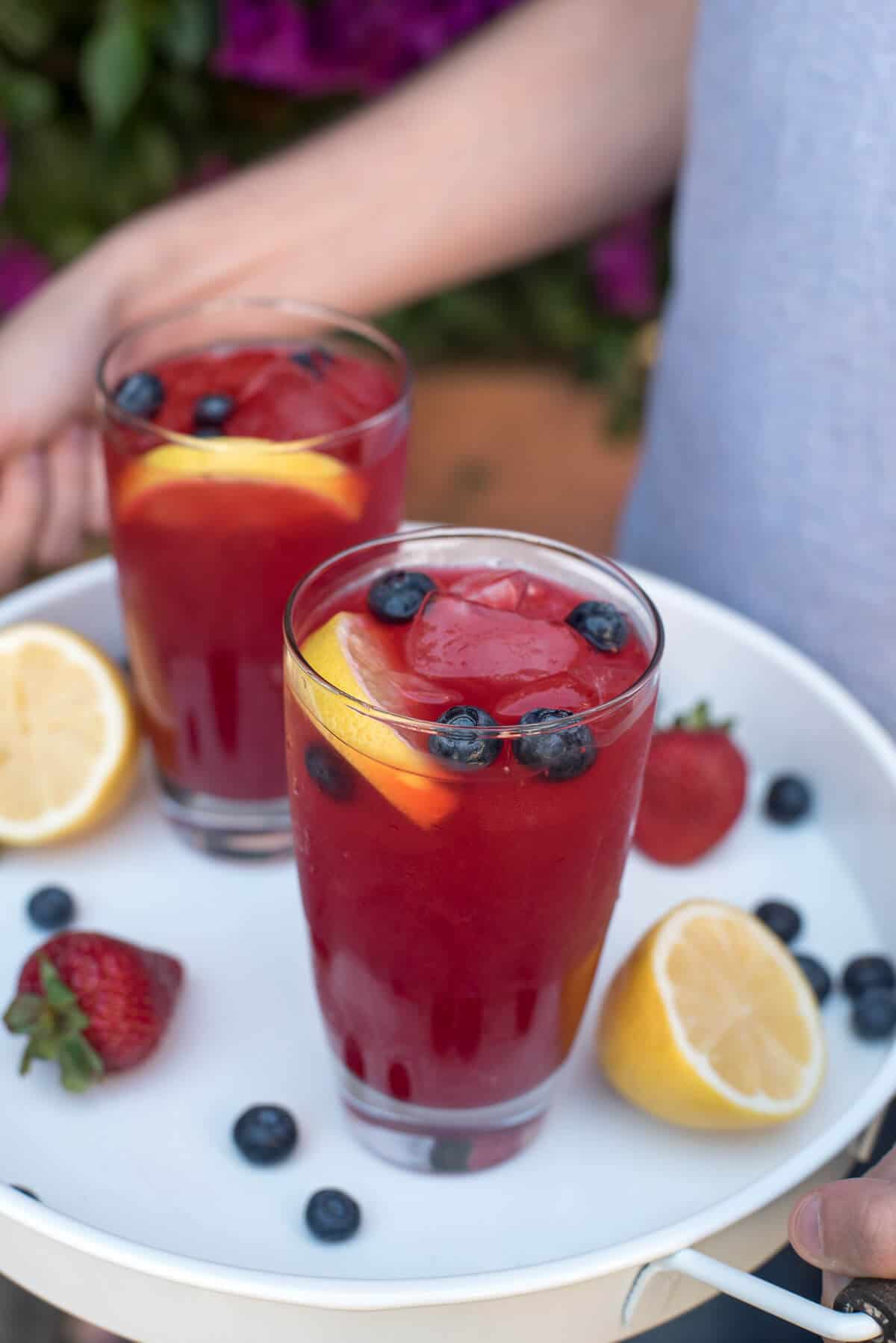 Hands holding a white tray with two glasses of Berry Lemonade.