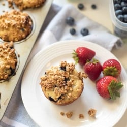 A blueberry muffin topped with granola on a white plate with strawberries.